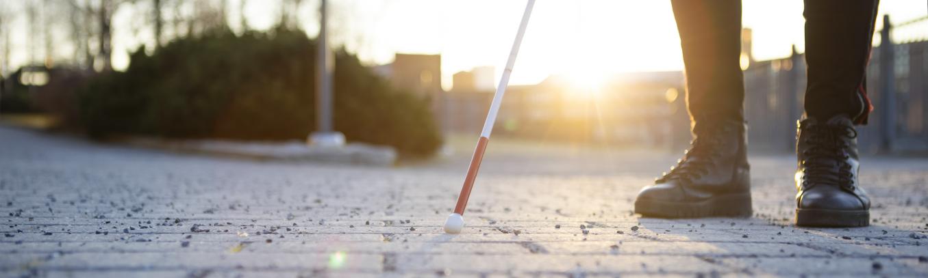 henkilö seisoo mukulakivikadulla, kuvassa jalat, valkoinen keppi ja auringon paiste 